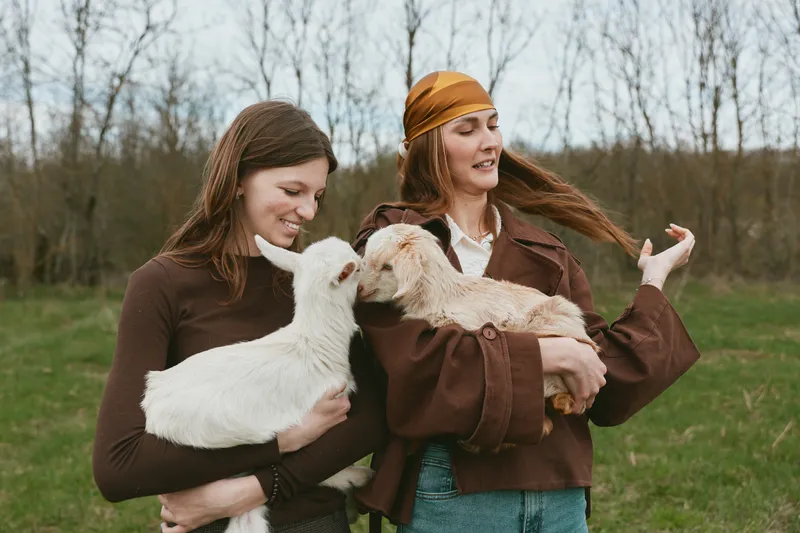 Two women outdoors in a rural field hold white goats, sharing a warm, playful moment. Casual jackets, natural light, and friendly livestock create a relaxed lifestyle scene.