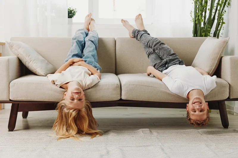 Two happy children lying upside down on a beige sofa in a bright living room, smiling and having fun. Concept of childhood joy, home, and playful moments.