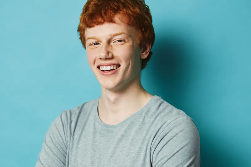 Young man with red hair and freckles wearing gray t shirt smiling with crossed arms on blue background. Happy male portrait with casual outfit and positive expression indoors.