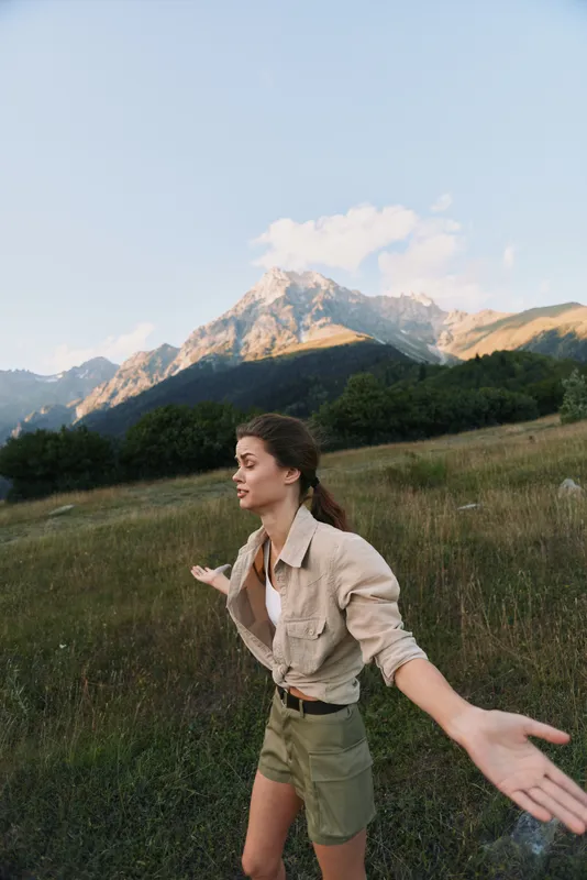 Young woman with outstretched arms enjoying nature in a mountain meadow during daytime. Concept of lifestyle, freedom, and outdoor adventure with clear sky background.