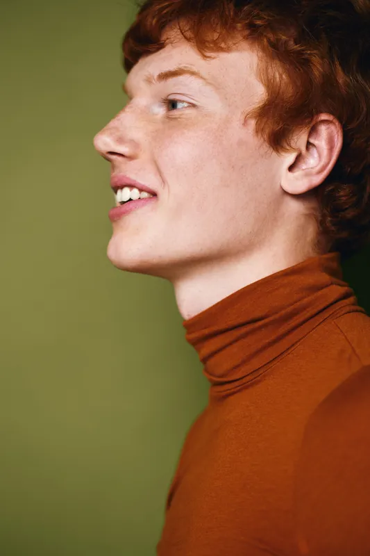 Profile of young man with red hair wearing brown turtleneck sweater against green background. Smiling male person with fair skin and casual clothing in studio portrait setting.