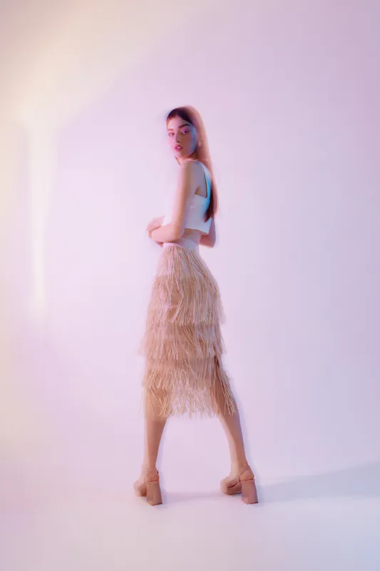 Young woman in elegant fashion outfit poses in soft studio lighting. She wears a fringed skirt and neutral top, showcasing beauty and style in a minimalistic setting.