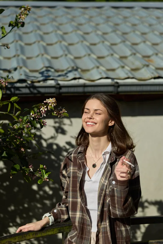 A smiling woman enjoys sunny outdoor moments in a plaid shirt, eyes closed, leaning on a railing in a garden. Concept: relaxed lifestyle and fresh air