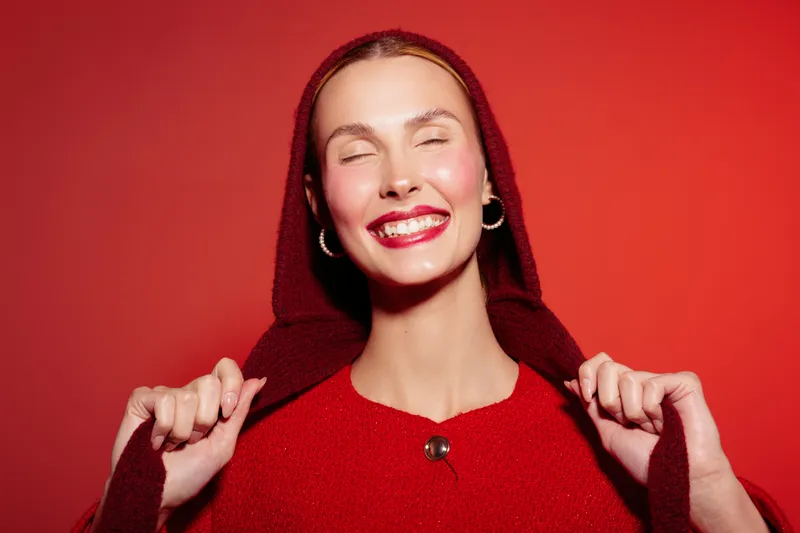 Smiling woman in red sweater and hood poses against red background with closed eyes and bright lipstick. Concept of happiness, fashion, and vibrant color contrast.