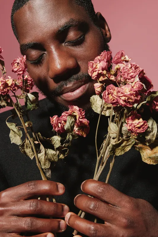 African man with dark skin holding dried pink flowers close to face in creative conceptual visual style with closed eyes and peaceful expression on pink background.