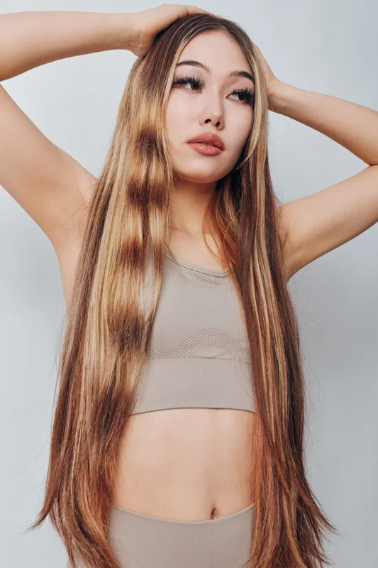 Woman with long straight hair posing with hands behind head against white background. Fashion model in beige outfit showing confident expression and natural makeup indoors.