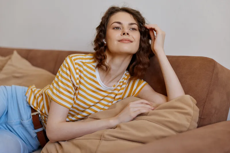 Casual woman lounging on a sofa at home, wearing a striped tshirt and jeans, relaxed pose and gentle smile. Concept: everyday comfort, home life, and tranquil mood.