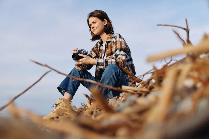 Casual photographer, woman in plaid shirt and jeans sits among driftwood, holding a camera, capturing outdoor nature and landscape.