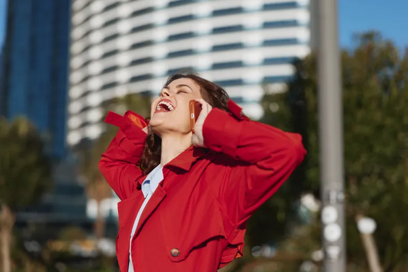 Happy woman in a red jacket talks on the phone outdoors, sunlight on her face, urban background, bright mood, candid city moment