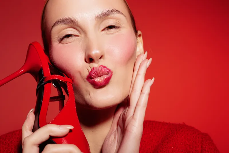 Close-up of a woman with red lipstick holding a red high heel shoe near her face on a red background. Concept of fashion, beauty, style, and glamour with vibrant colors.
