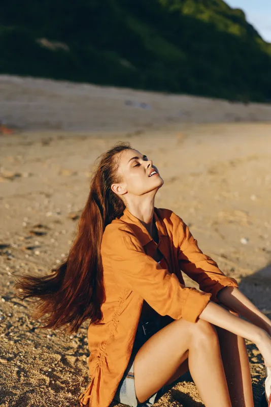 A beach scene with a woman sitting in warm sand, eyes closed, face uplifted to the sun. Relaxed pose, light wind through hair, orange shirt, calm mood, shoreline backdrop.