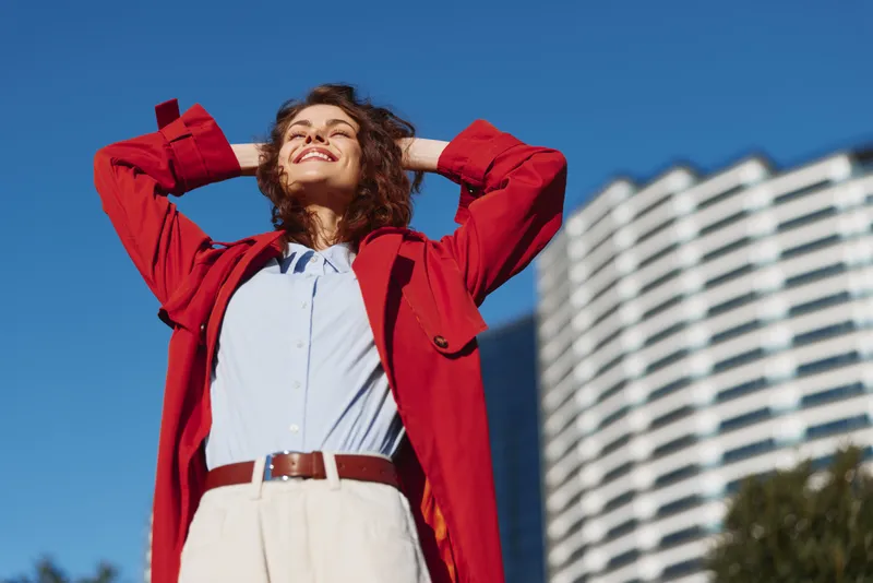 Young woman in a red coat enjoys a sunny day outdoors with her hands behind her head and eyes closed, standing in front of a modern white building under a clear blue sky.