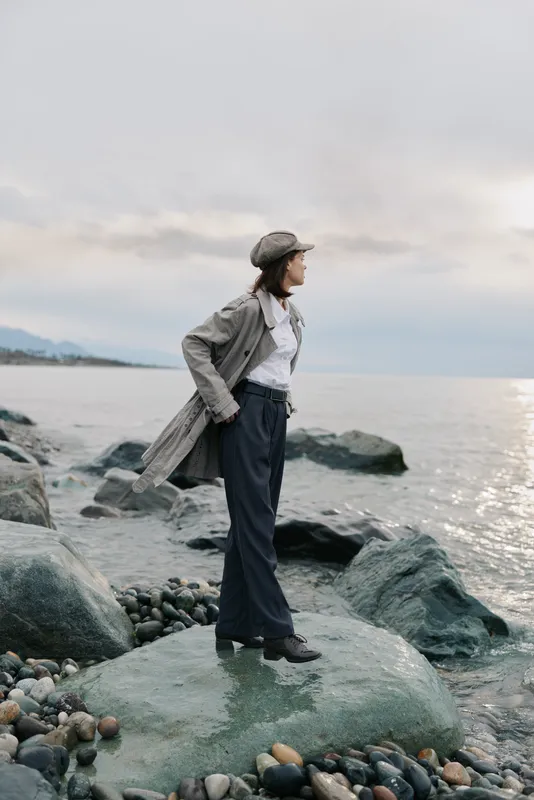 Woman in stylish coat and hat stands on rocky shore looking at calm sea under cloudy sky. Concept of autumn fashion, nature, and peaceful outdoor moment.