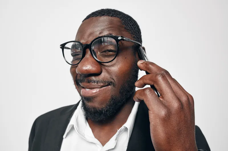 African American man with dark skin wearing glasses and suit jacket smiling while talking on smartphone against white background. Closeup portrait of confident businessman.