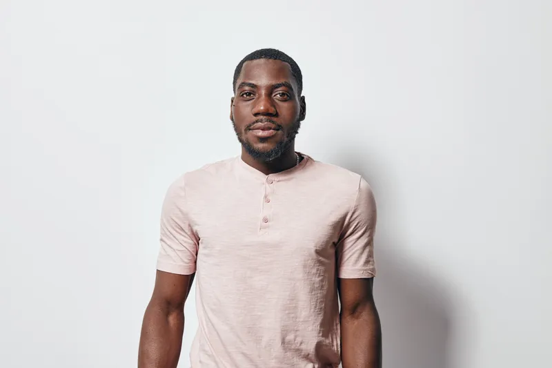 African American man with dark skin wearing light pink shirt standing against white background looking at camera with neutral expression and short hair.