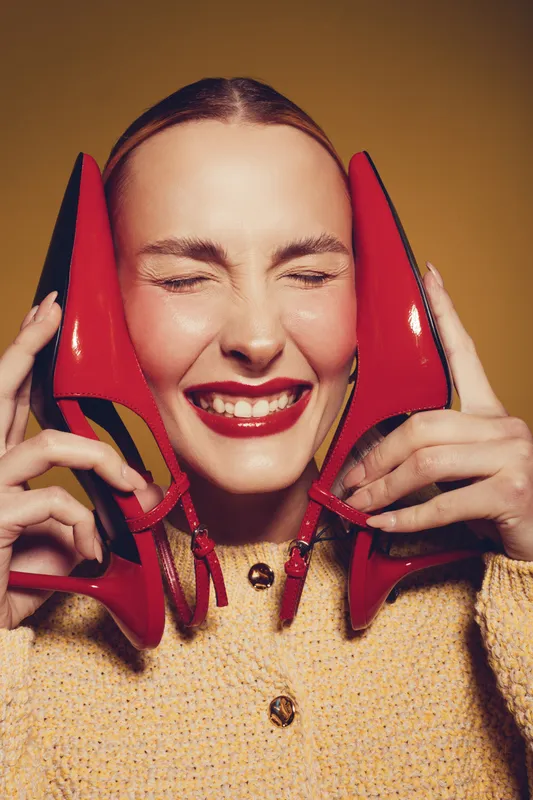 Happy woman with red lipstick holds red high heels near face, eyes closed and smiling. Fashion concept with bright colors and joyful mood on warm background.