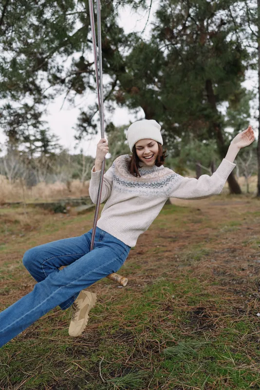 Woman on a wooden swing in a park, wearing a cozy sweater and beanie, enjoying a carefree moment in nature with trees and open space around.