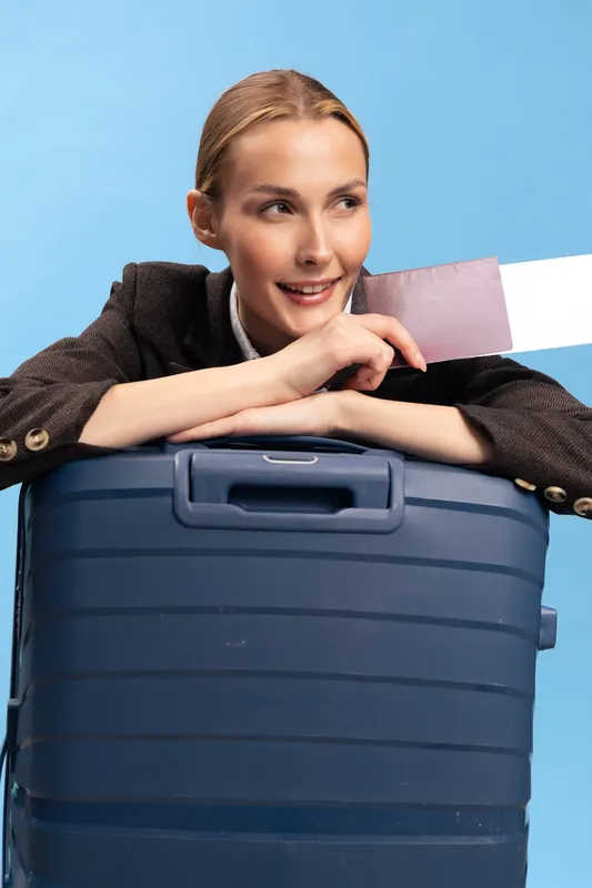 Smiling woman with light hair leaning on large blue suitcase holds boarding pass, looking away. Concept of travel, vacation, and holiday preparation in studio with blue background.