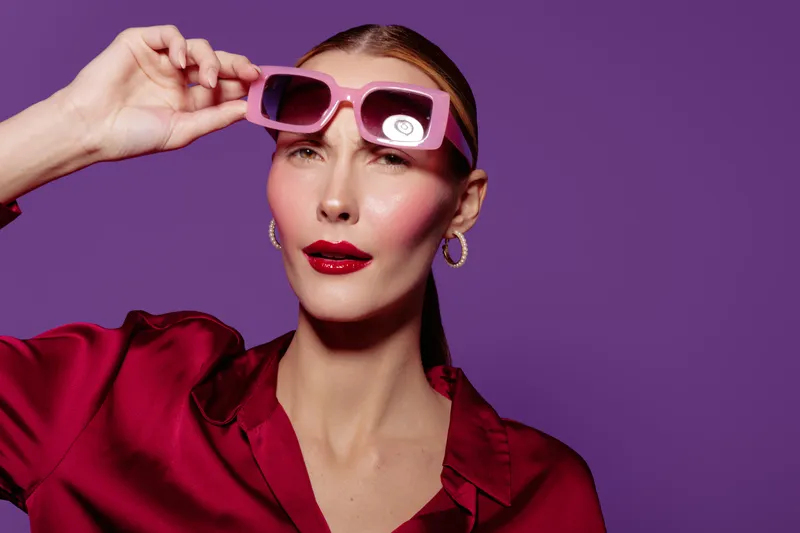 Stylish woman in red blouse adjusts pink sunglasses on purple background. Fashion portrait with bold makeup and confident expression. Concept of style, beauty, and modern accessories.