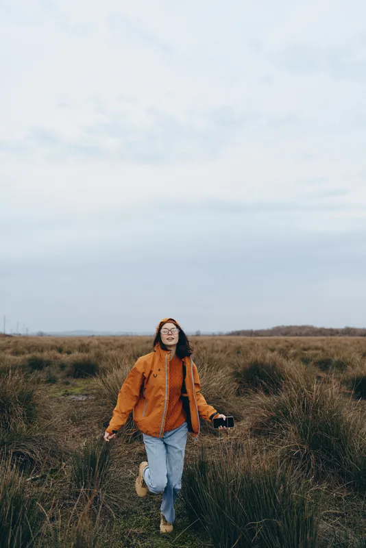Woman lifestyle in an outdoor field, walking through tall grass in orange jacket under cloudy sky, relaxed travel mood, nature scenery, countryside horizon, solo, open air, wide landscape.