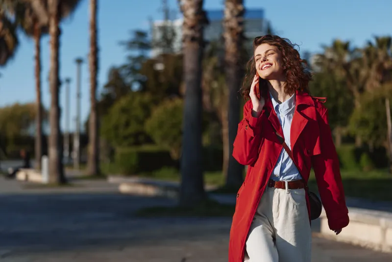 Smiling woman with curly hair wearing a red coat walking outdoors on a sunny day. Casual fashion mood in a palm tree lined avenue, bright and energetic.