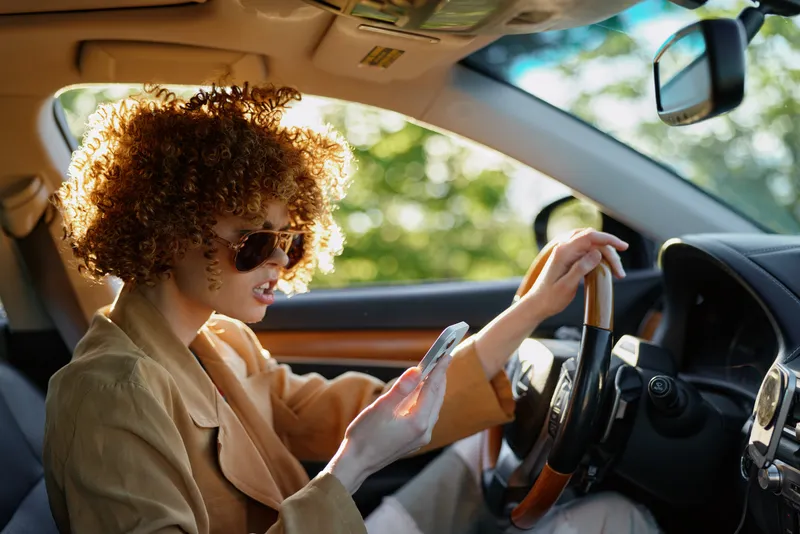 Young woman with curly hair drives a car wearing a tan coat and sunglasses, enjoying a sunny daytime road moment behind the wheel.