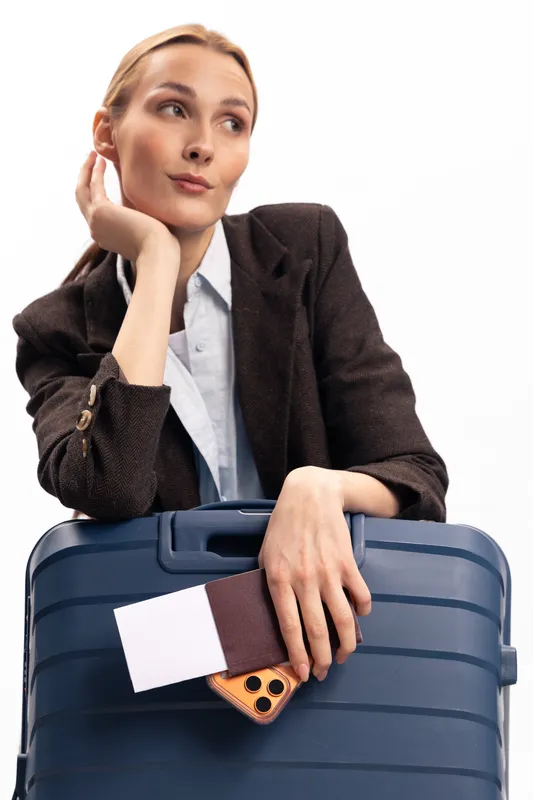 Young woman in black jacket holds blue suitcase and phone with ticket, looking away with thoughtful expression on white background. Concept of travel, business trip, and planning.