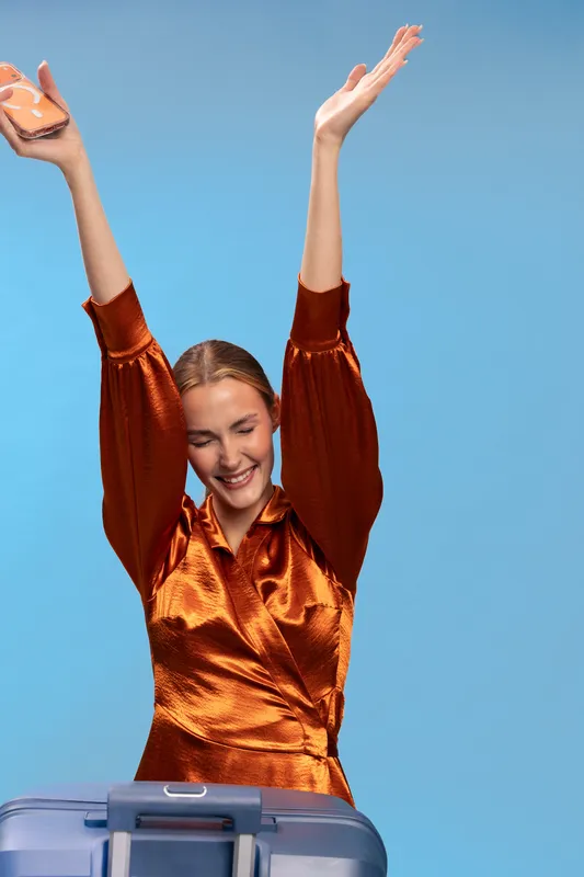 Happy woman in shiny orange dress raises hands celebrating near suitcase on blue background. Concept of travel excitement, vacation, and joyful journey with positive emotions and freedom.