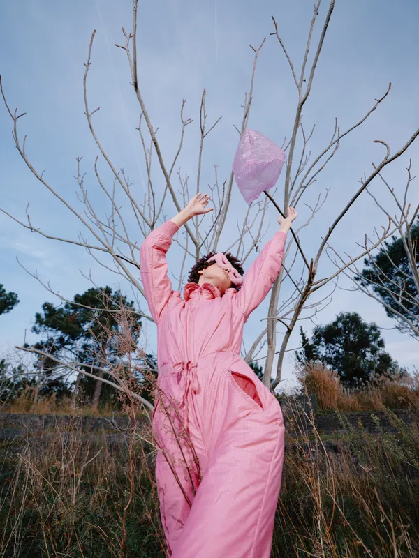 Woman in a pink dress outdoors releases a paper lantern beside leafless trees under a blue sky, capturing a joyful moment of celebration and wind as imagination lifts.