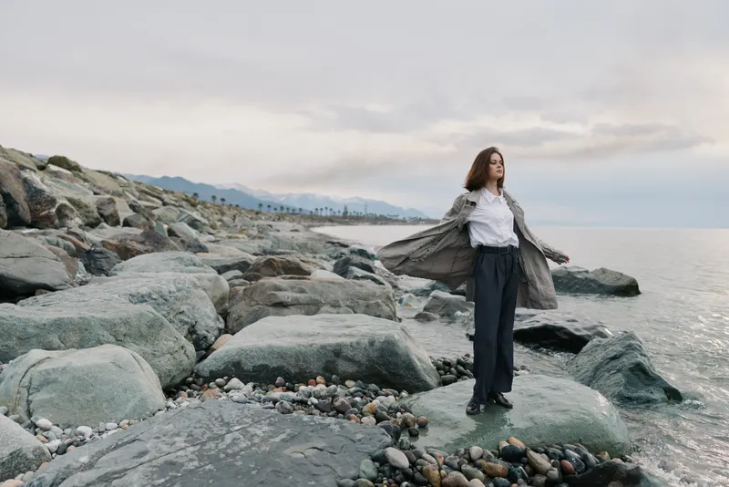 Young woman stands on rocky beach wearing coat and white shirt. Calm sea and cloudy sky in background create peaceful atmosphere. Concept for freedom, nature, and contemplation.