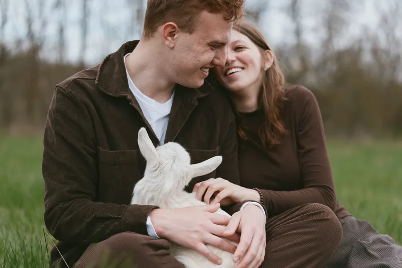 Young couple sitting on grass in casual clothes enjoy a tender moment while holding a baby goat in a natural outdoor setting during early spring.