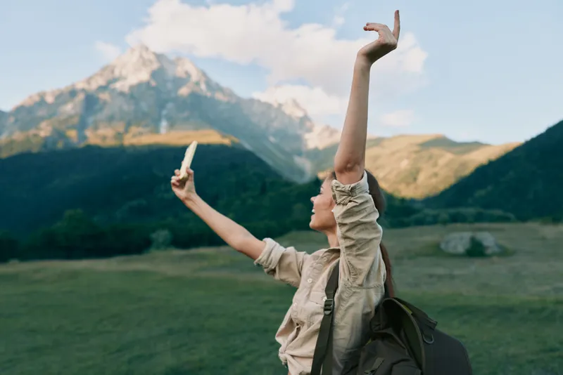 An outdoorsy woman in a beige shirt raises her phone in a mountain meadow, celebrating exploration and freedom. A vivid lifestyle moment of travel and adventure.