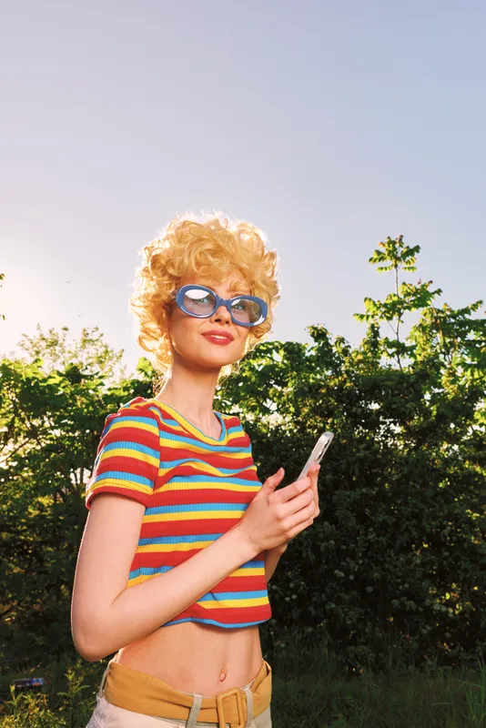 Woman in striped retro top and sunglasses stands outdoors holding a smartphone, capturing casual sunny moments with vintage film color and a carefree mood, bright laughter