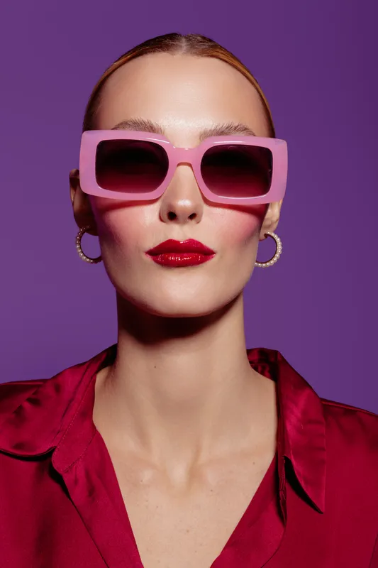 Stylish woman with slicked hair wears oversized pink sunglasses and red lipstick. She poses confidently against a purple background wearing a red satin blouse and gold hoop earrings.
