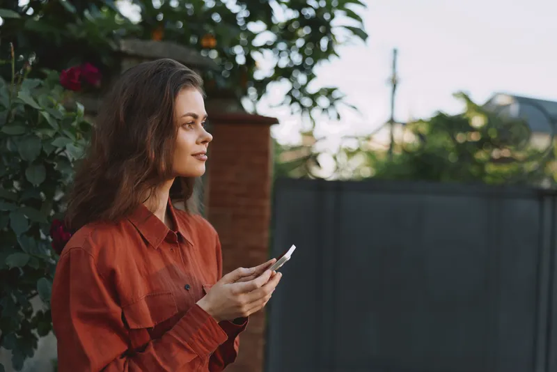 A young woman in a red shirt stands outdoors, using a smartphone with a thoughtful expression, enjoying a quiet moment in a garden