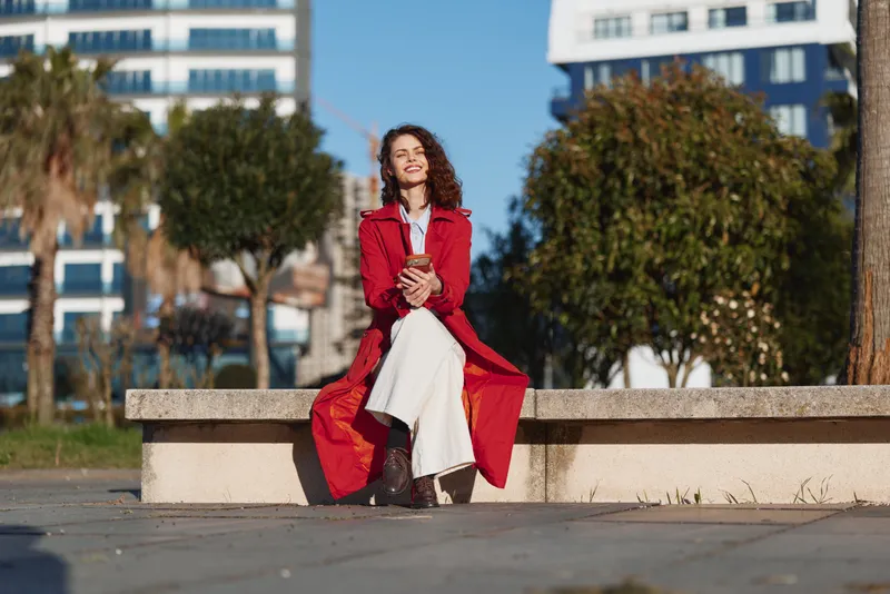 Young woman in a red coat sits on a stone bench outdoors holding a smartphone and smiling with closed eyes on a sunny day.
