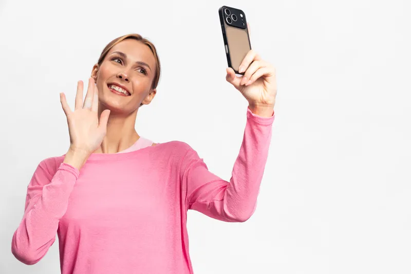 Smiling woman in pink sweater takes selfie with smartphone and waves hand. Isolated on white background. Concept of communication, technology, and social media interaction.