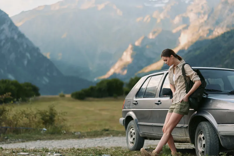 A young woman in casual clothes leans against a car on a rural road with mountains in the background. The scene reflects outdoor lifestyle and travel adventure.