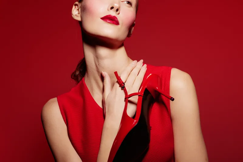 Stylish woman in red dress holds red high heel shoe near neck on red background. Fashion concept with bold makeup and elegant pose in studio lighting for glamour and style.
