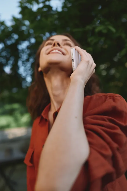 Bright outdoor portrait of a smiling woman talking on a phone, looking up at the sky, candid lifestyle moment, warm sunlight, relaxed posture, natural colors, leafy background