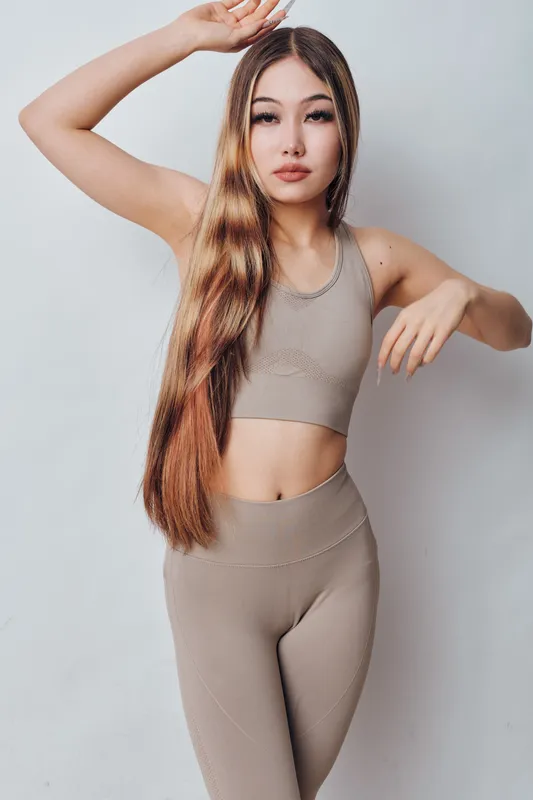 Young woman with long hair wearing beige activewear posing with hand raised near head and other extended, fitness outfit for health and exercise lifestyle indoors.