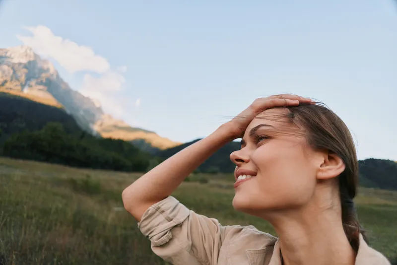 A sunlit young woman in a field shields her eyes with a hand as mountains rise in the background, capturing a candid outdoor moment of relaxed confidence.