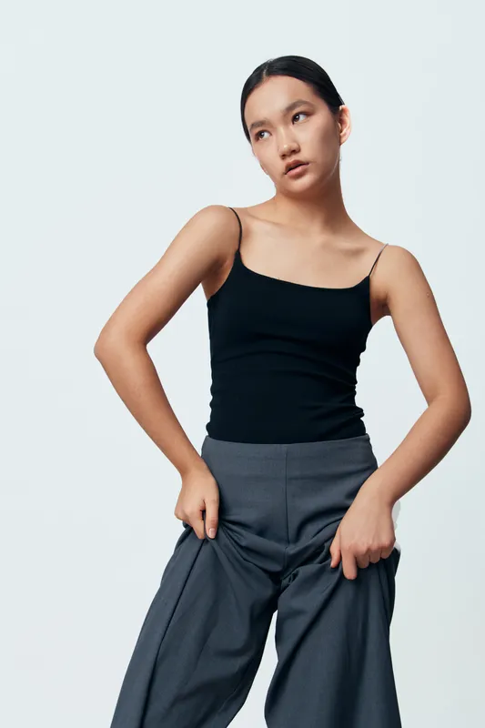 Young woman wearing black tank top and gray pants posing with hands in pockets against white background in studio for fashion or casual style portrait photo.