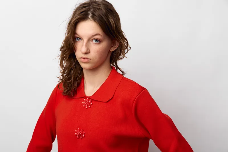 Young woman with wavy brown hair wearing a red sweater with decorative buttons looks seriously at camera on white background. Concept of fashion, portrait, and attitude.