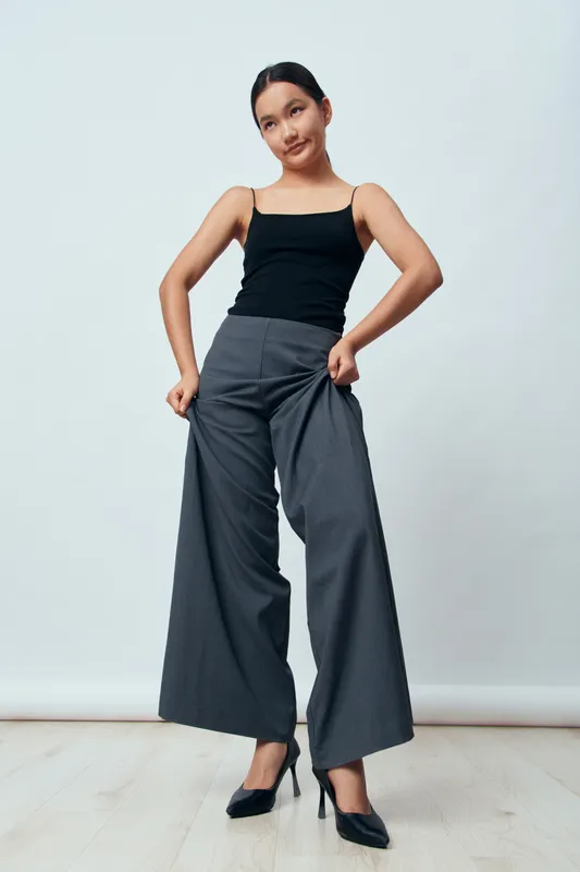 Woman model posing in wide leg gray pants and black tank top with pointed high heels on light floor with plain background studio fashion outfit casual style confidence