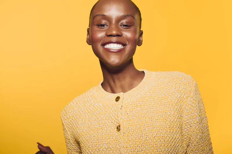 Smiling black woman with dark skin wearing a yellow knitted sweater poses against a bright yellow background. The image conveys happiness, confidence, and vibrant style.