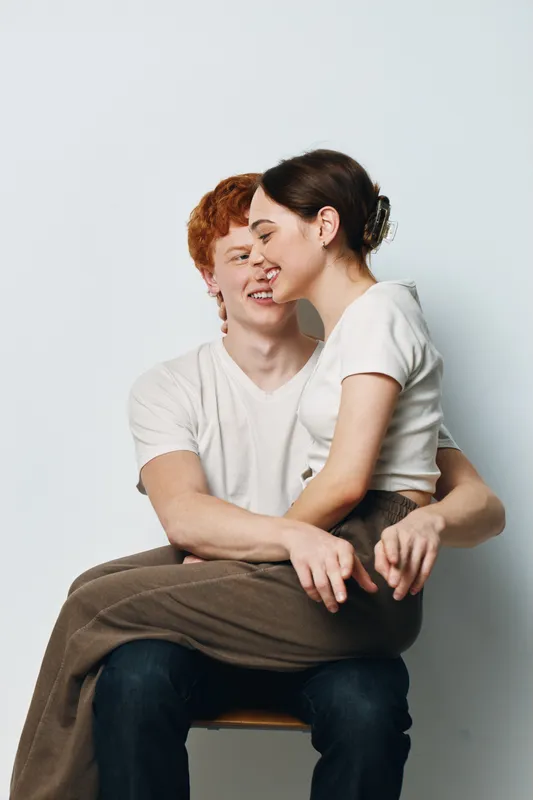 Couple sitting and hugging on chair with smiles and affection in studio setting. Young man and woman in casual clothes showing love and connection closeup.