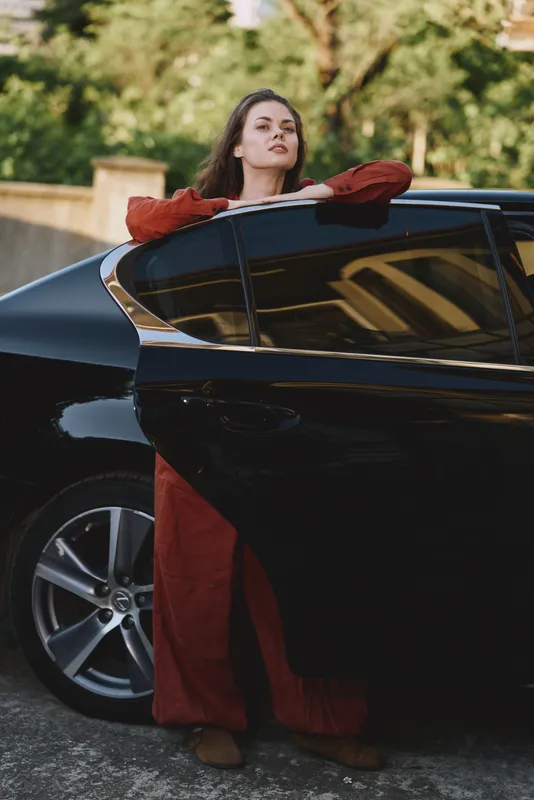 One young woman leans on a sleek black car outdoors, wearing a red dress with long sleeves, exuding confidence and a relaxed, stylish vibe