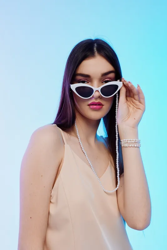 Young woman in beige dress wearing white sunglasses and pearl accessories poses against blue background. Concept of beauty, fashion, and stylish accessories.