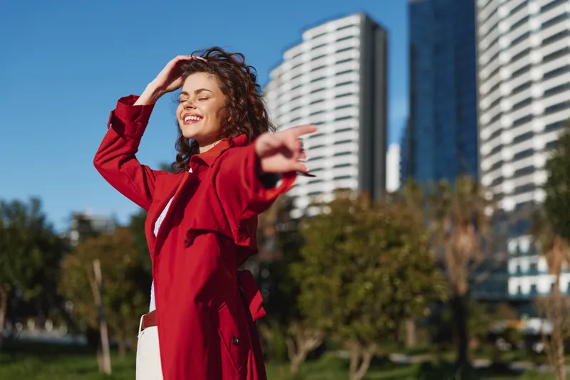 Young woman in a red coat enjoys the sunny day outdoors with her eyes closed and a joyful smile, surrounded by greenery and modern buildings in the background.